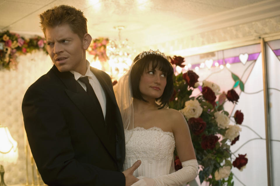 A bride in a strapless white gown and tiara stands beside a groom in a black tuxedo, looking concerned, in a decorated wedding venue with roses and heart motifs