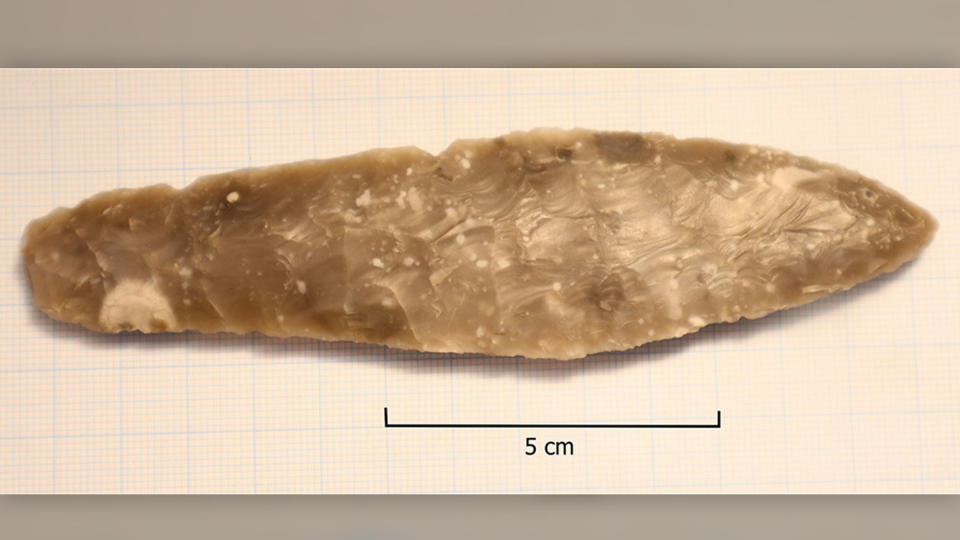 a light brown and grey piece of carved stone sitting on a white table. The stone is shaped like a very squat dagger