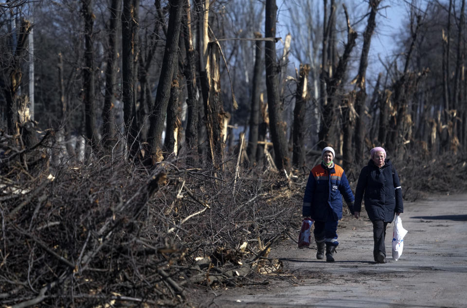 Two people wearing coats and hats and carrying handbags walk hand-in-hand along a roadway lined with splintered trees and fallen branches.