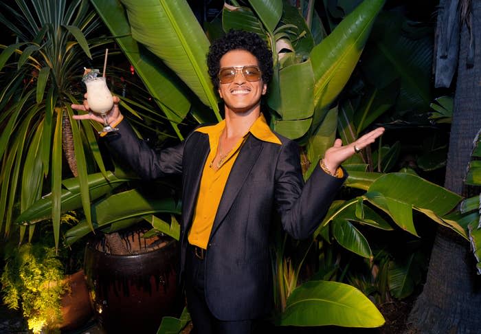 Bruno Mars posing with a drink, sporting a black blazer over a yellow shirt. He's smiling against a backdrop of tropical plants