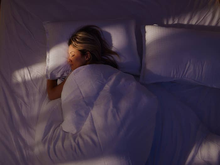 A person is lying in bed sleeping under a blanket, with their head resting on a pillow. Sunlight casts shadows on the bedding