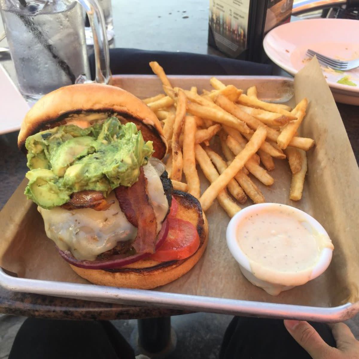 BJ's Brewhouse Bacon-Guacamole Deluxe Burger with fries and a cup of garlic parmesan sauce on brown serving paper and serving tray with glass of water and other items in the background