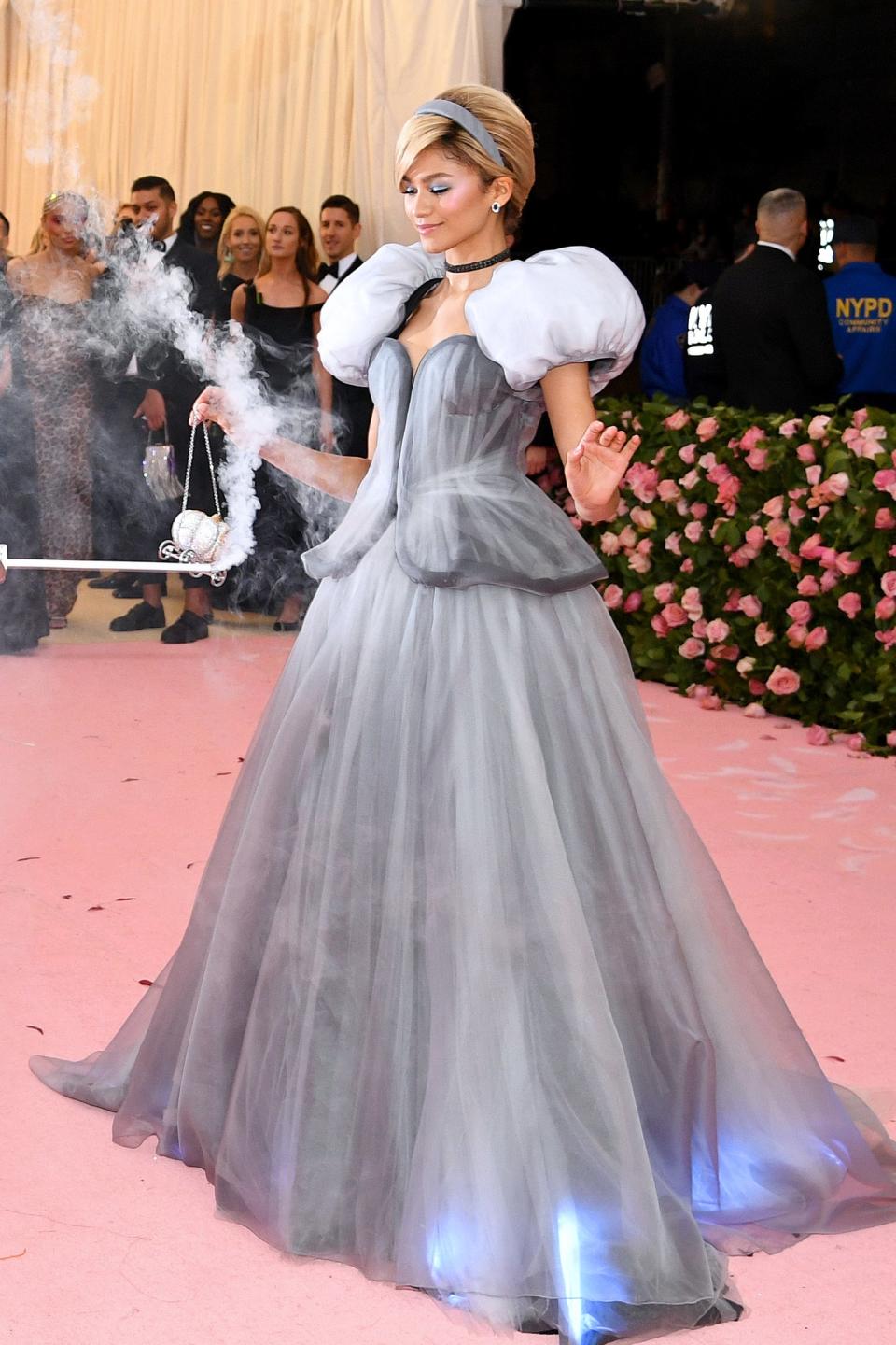 Zendaya in a light grey gown with a lit-up glass slipper, person in a blue costume with a wand
