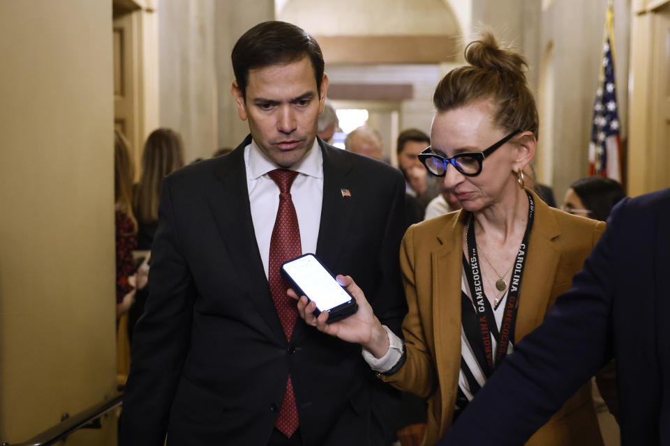 Sen. Marco Rubio walks down a corridor in the Capitol talking, as a reporter holds up her cellphone to record him.
