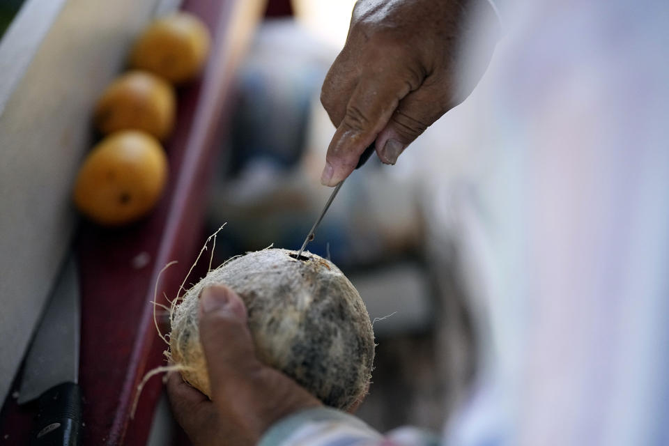 Andres Matamoros prepares a cold coconut for a customer while selling fresh fruit from his roadside stand Wednesday, June 28, 2023, in Houston. Meteorologists say scorching temperatures brought on by a heat dome have taxed the Texas power grid and threaten to bring record highs to the state. (AP Photo/David J. Phillip)