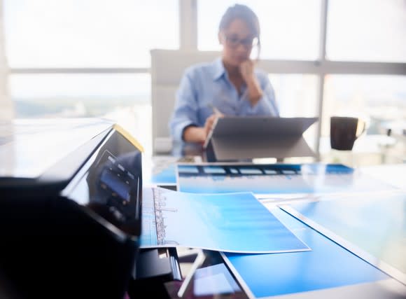 Printer prints out color displays while woman works on laptop in background.