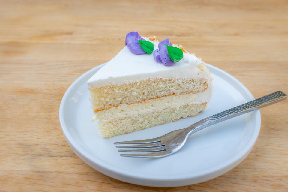 Slice of vanilla cake with layer of vanilla filling and white icing and purple rose buds on top next to a fork