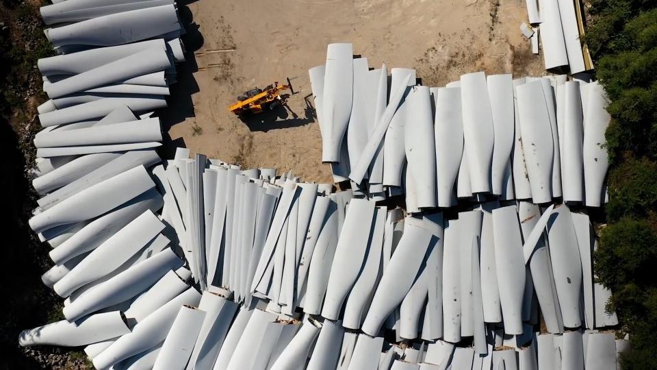 Wind turbine blades piled up outdoors on sandy ground.