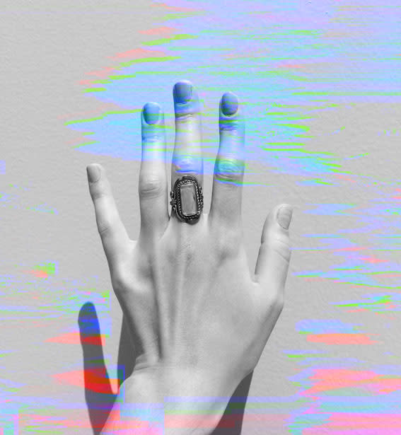 A hand with neatly manicured nails showing an ornate ring on the middle finger against a glitchy, distorted background. No text or identifiable people are present