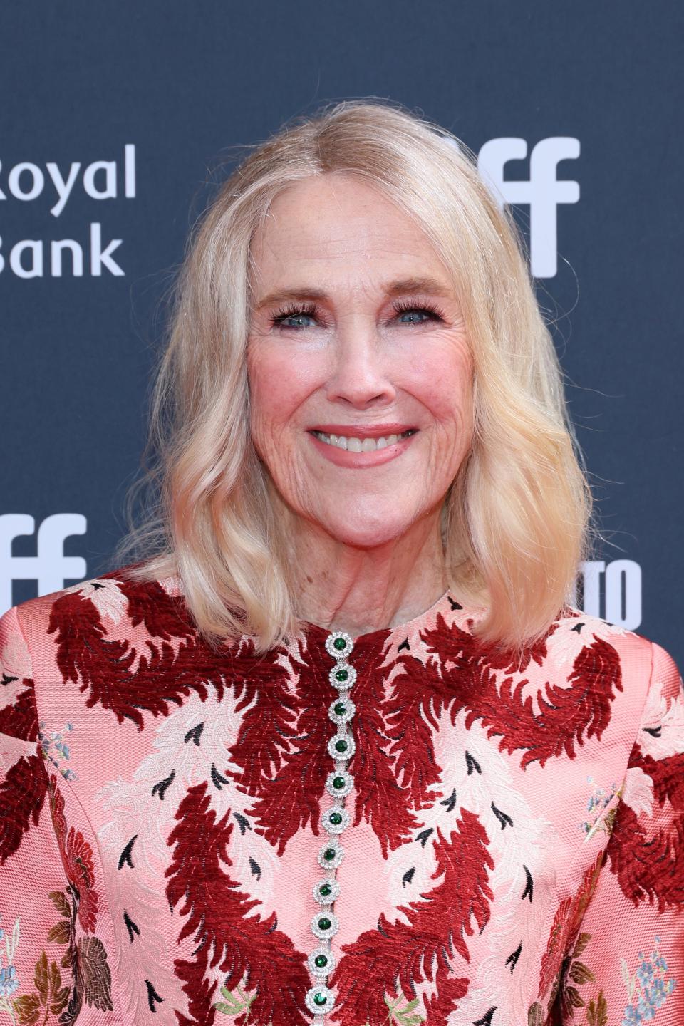 Catherine O'Hara smiles for the camera wearing a floral dress with intricate red details at a film festival event.