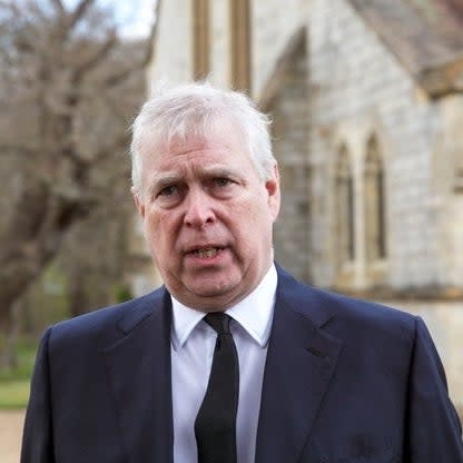 Prince Andrew has a serious expression and stands outside a stone building, wearing a black suit and tie