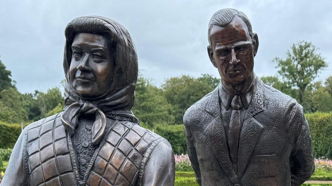 The bronze sculpture of the royal couple and their two corgis in the Parterre Garden at Antrim Castle Gardens, surrounded by flowers and under a grey looking sky with trees in the background. 

The late queen (sculpture) is wearing a cardigan, scarf and carrying a handbag. She is holding her hands together. 

The late king (sculpture) is wearing a suit. 

The corgi dogs are sitting down, one is looking up at the queen and one is looking ahead. 