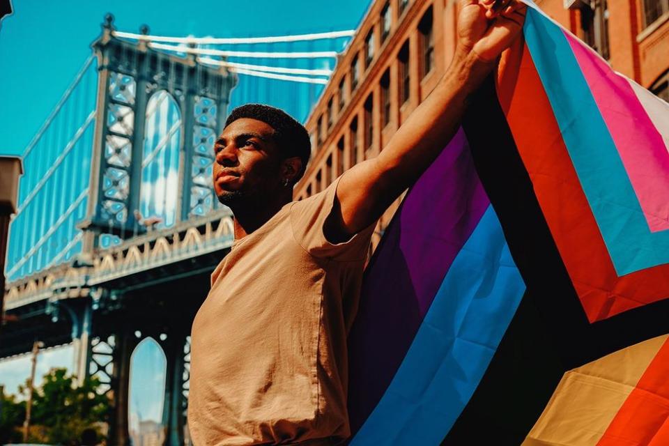 Young black man with arms outstretched holding Progress Pride flag with Brooklyn Bridge in background