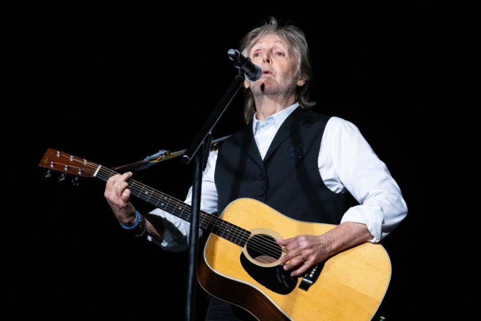 paul mccartney, wearing a black vest and white shirt, sings into a microphone while playing guitar, standing in front of a black backdrop