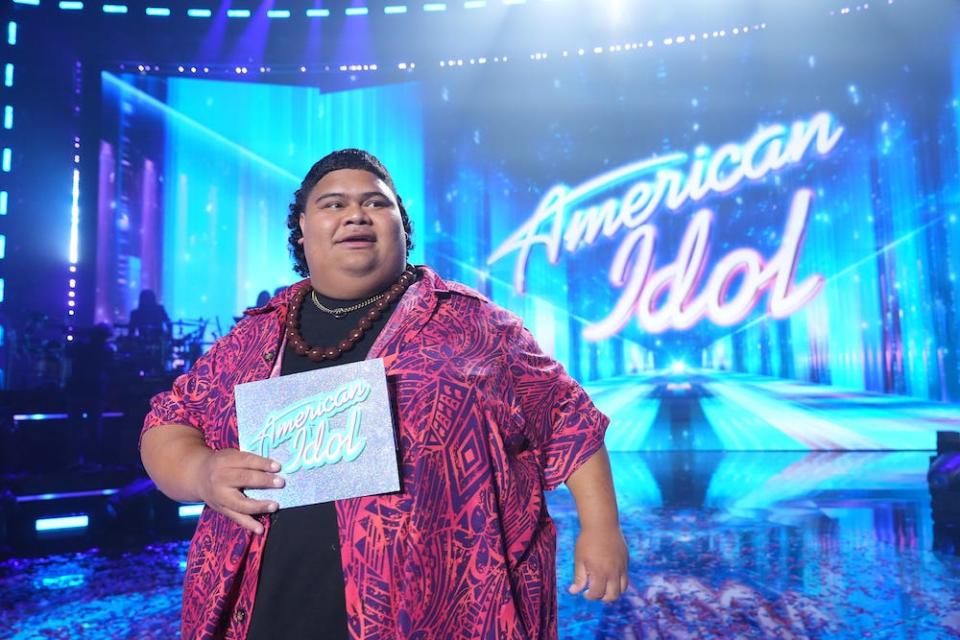 Hawaiian artist Iam Tongi stands on the bright blue set of American Idol, with the show's name behind him, with a smile on his face, in an undated image.