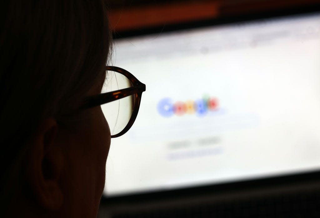 A shadowy person wearing glasses sits in front of a blurry laptop screen displaying the Google search engine.