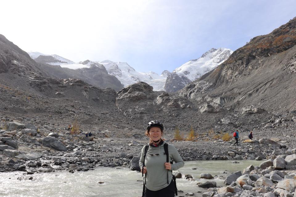 woman wearing bike helmet standing in front of pale green creek under rocky mountains slopes with a glacier behind