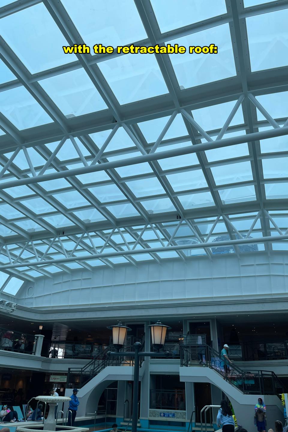 Glass ceiling structure of a building with geometric patterns, text overlay mentioning a retractable roof. People visible below