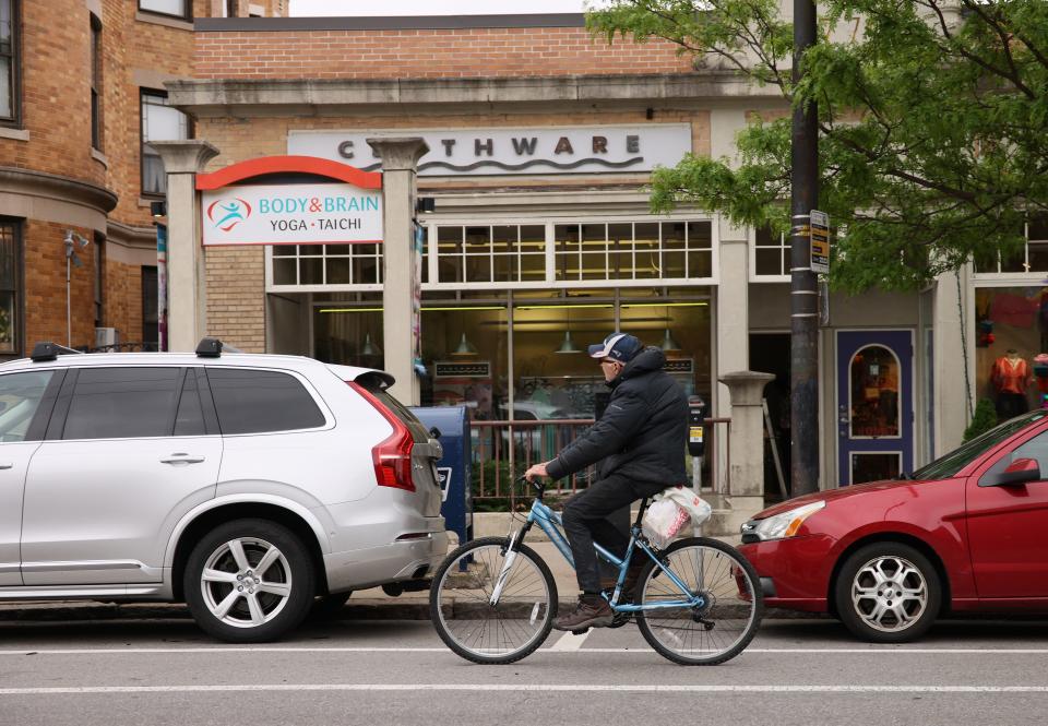 An older man in a baseball cap and parka rides a blue bicycle on a city shopping street, past parked cars and a yoga studio