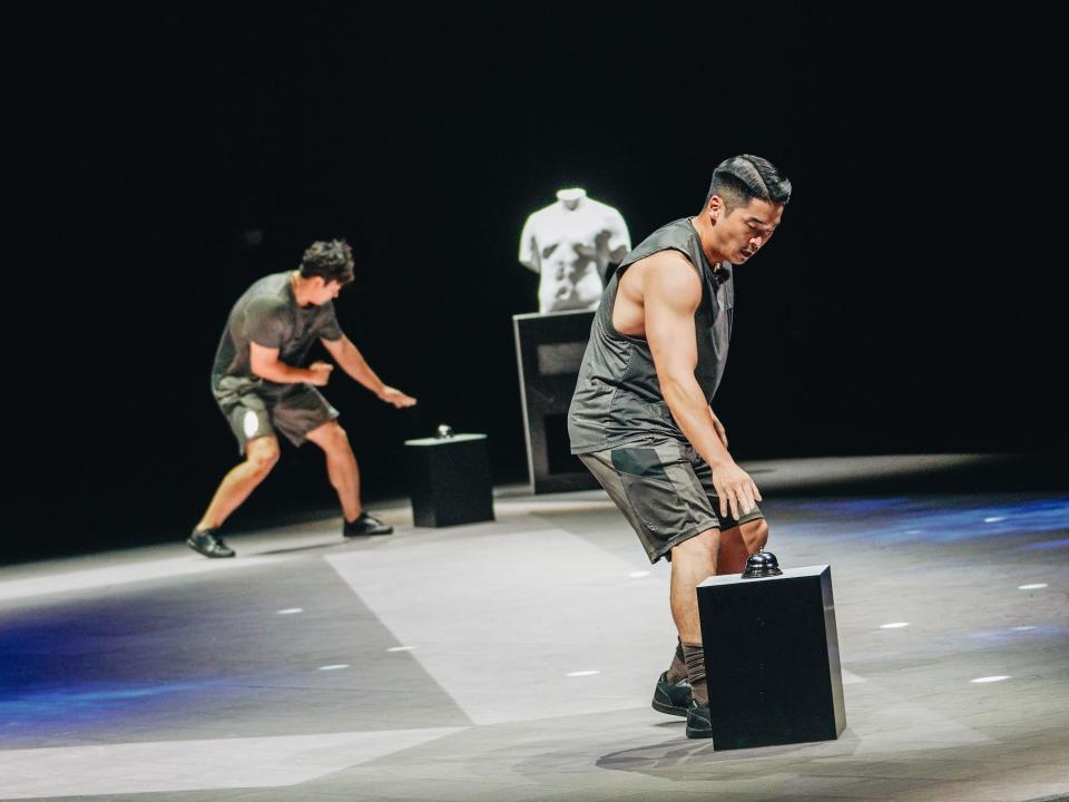 woo jin-yong on physical 100, his hand above a bell that he has to ring in order to complete a challenge. behind him, park jin-yong is visible, standing in a similar stance and bending down to touch a bell