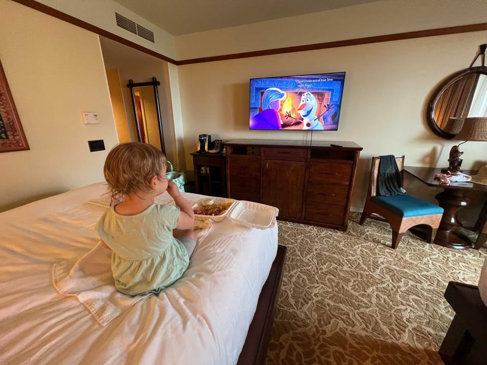 A child sitting on a hotel bed and watching TV while eating food.