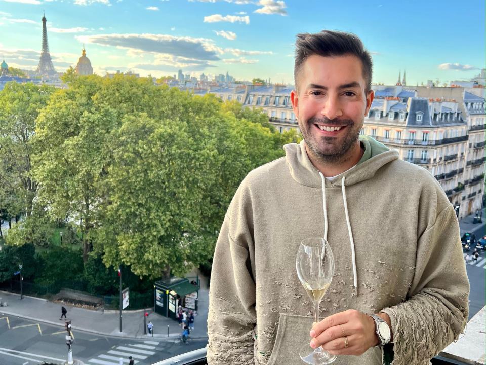 A man standing on a balcony with a view of the Eiffel Tower in the background, holding a glass of champagne.