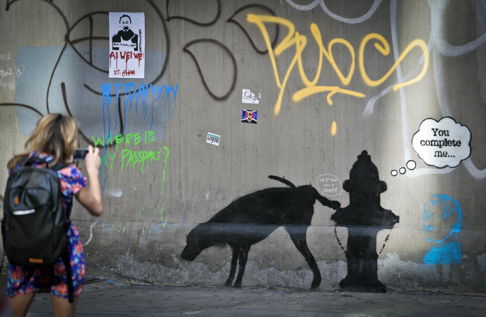 Graffiti by the secretive British artist Banksy, featuring a dog and a fire plug, draws attention on 24th Street, near Sixth Avenue in New York, on Friday, Oct. 4, 2013. Banksy graffiti is turning up on the streets of the city and all over social media. Banksy announced on his website that he is undertaking “an artists residency on the streets of New York” this month. (AP Photo/Bebeto Matthews)