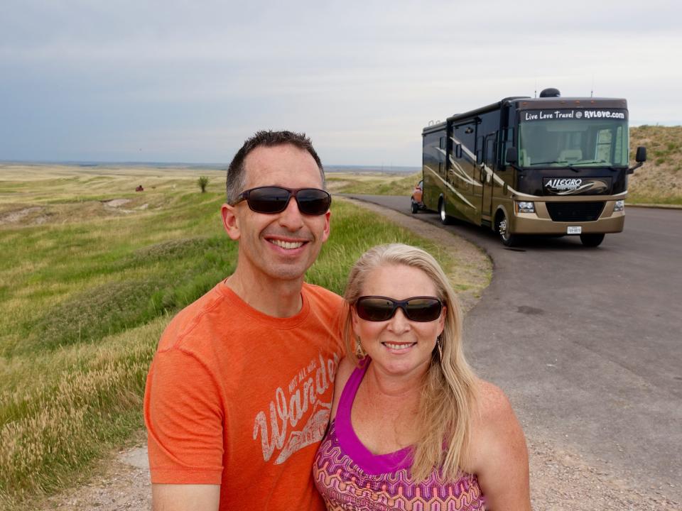 A couple with sunglasses on pose for a picture in front of their RV with green grass and cloudy skies behind them.