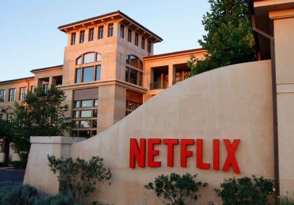 A photograph of the Netflix headquarters campus in Los Gatos, California, featuring a large Netflix logo on a wall in the foreground.