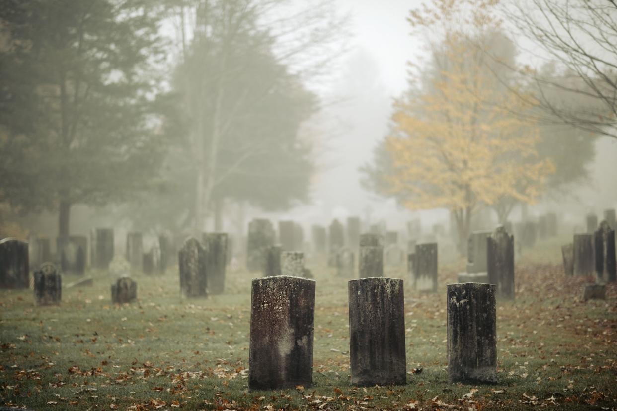 Three gravestones standing in a foggy graveyard during the fall. Focus is on front headstone.