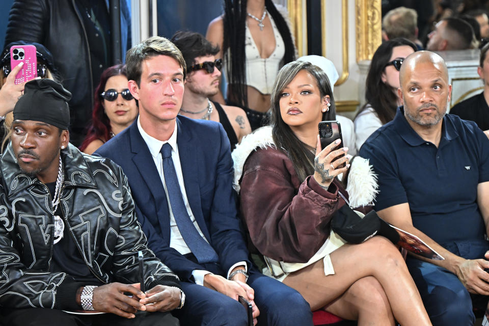 Pusha T, Paul Bourelly, and Rihanna sit in the front row at a fashion event, with Rihanna taking a photo
