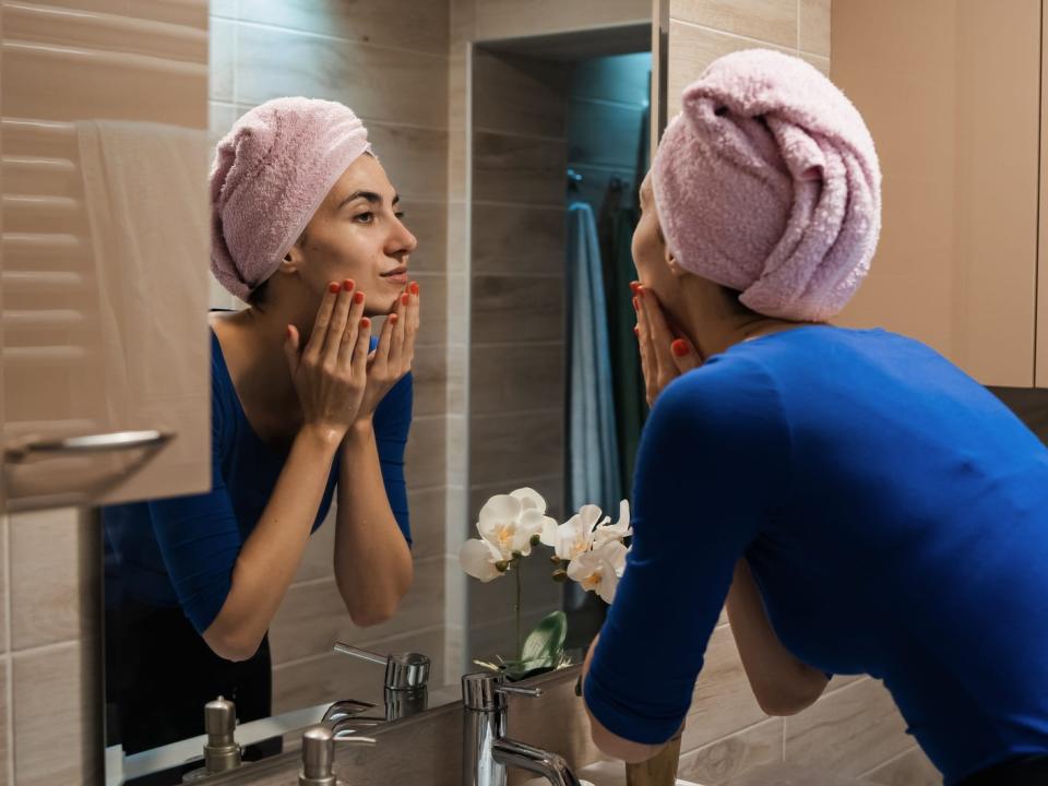 A woman wearing a towel on her head washing her face in front of the mirror