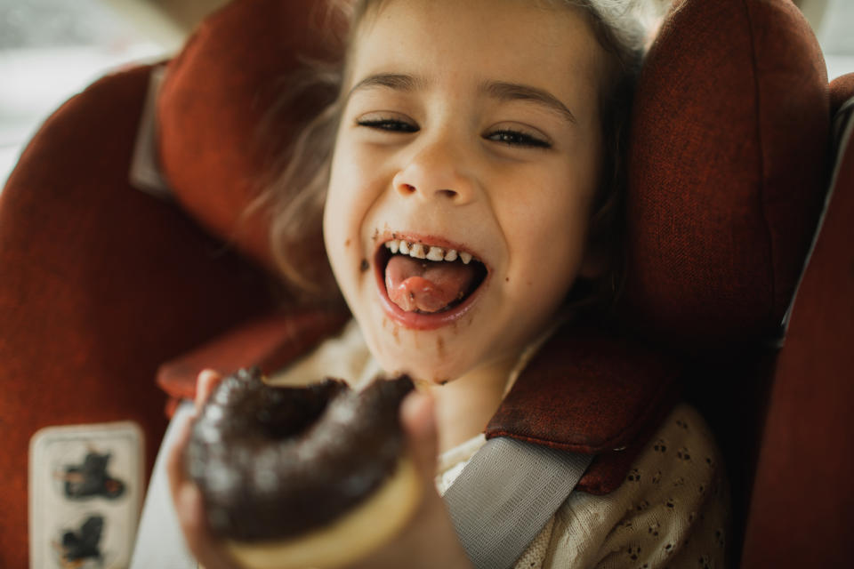 A child in a car seat smiles with chocolate on their face and holds a half-eaten donut