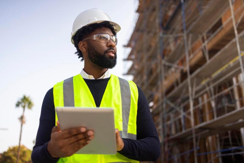 A man in a dark sweater, a reflective vest, and a white hard hat holds a tablet while standing in front of a building under construction.