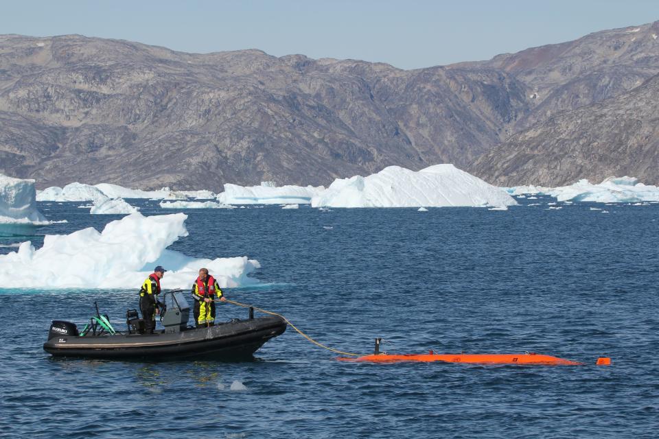 Two people on a dingy in the ocean holding a rope tied to the Ran submersible with ice and mountains in the background