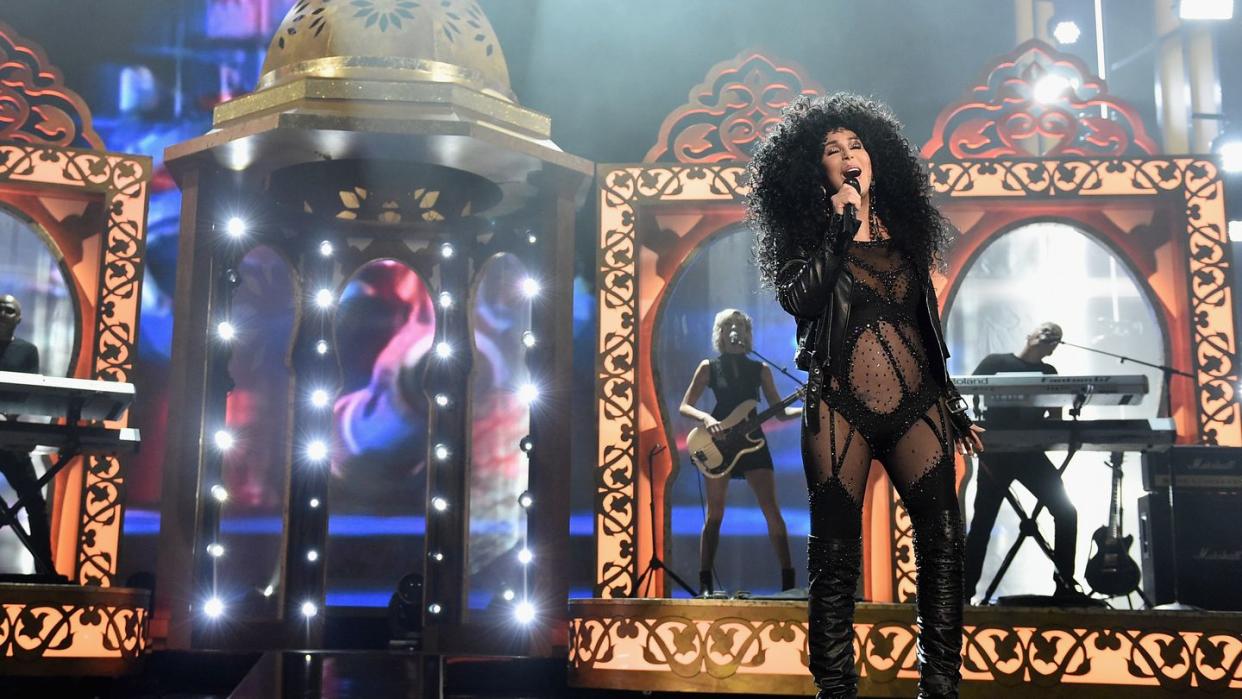 cher sings into a microphone she holds in one hand, she stands on a stage with props and musicians playing behind her