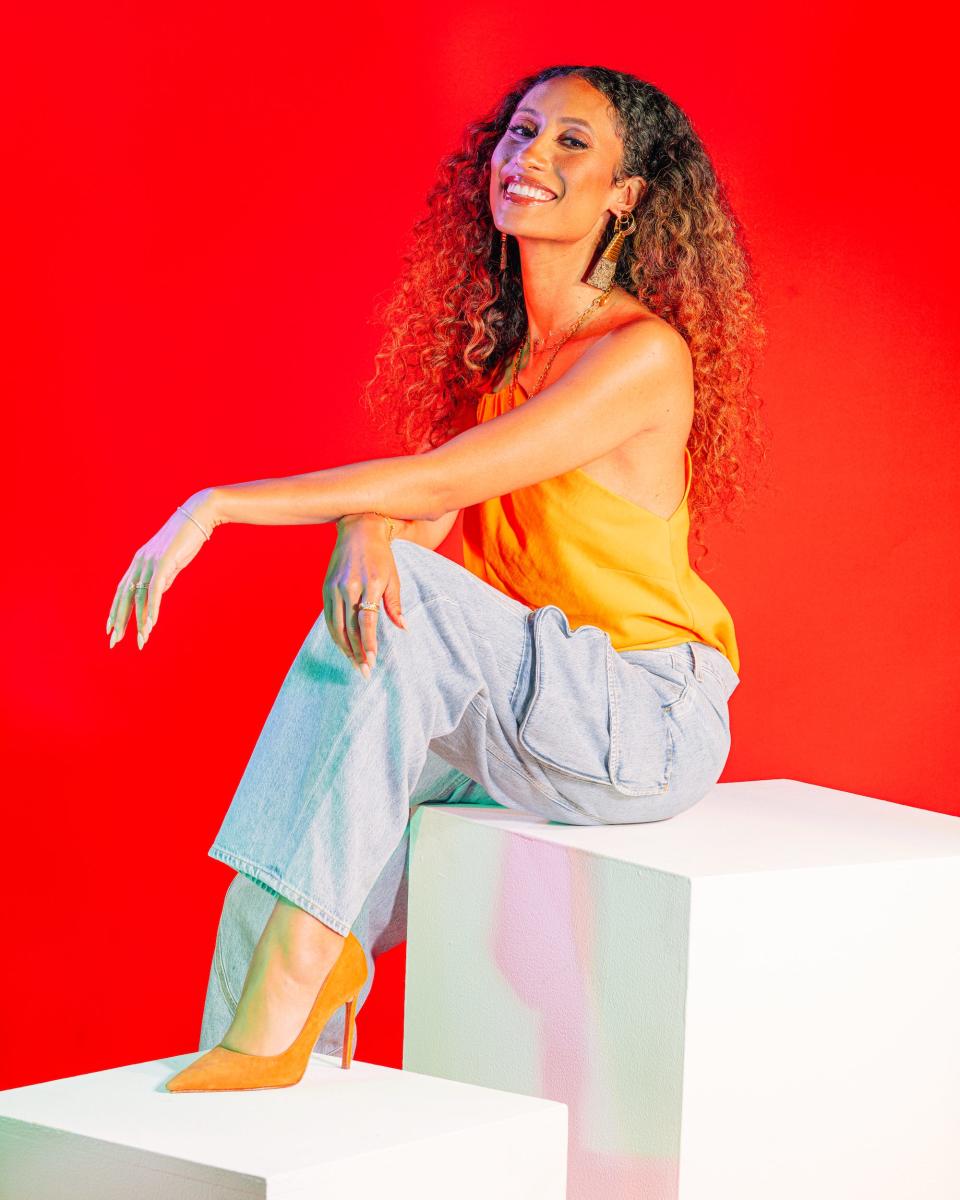 Elaine Welteroth in orange sleeveless top and blue cargo pants sits in front of a bright red studio background