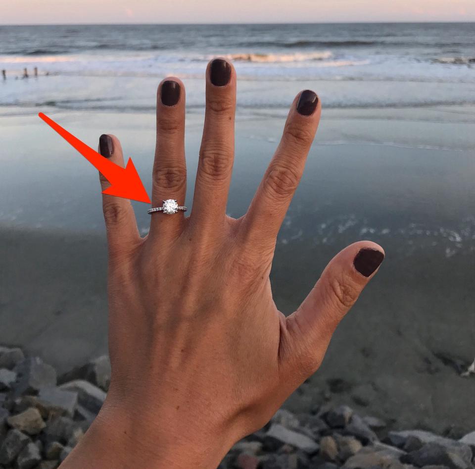 A red arrow points to an engagement ring on a woman's hand as she holds it up in front of the ocean.