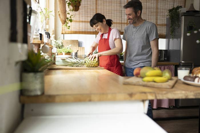 Happy mature couple at home kitchen preparing food, cutting pineapple, domestic life