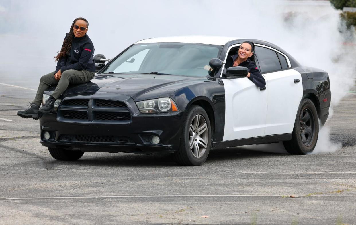 One woman sits on the hood of a fake cop car while another woman smiles and leans out the driver's-side window.