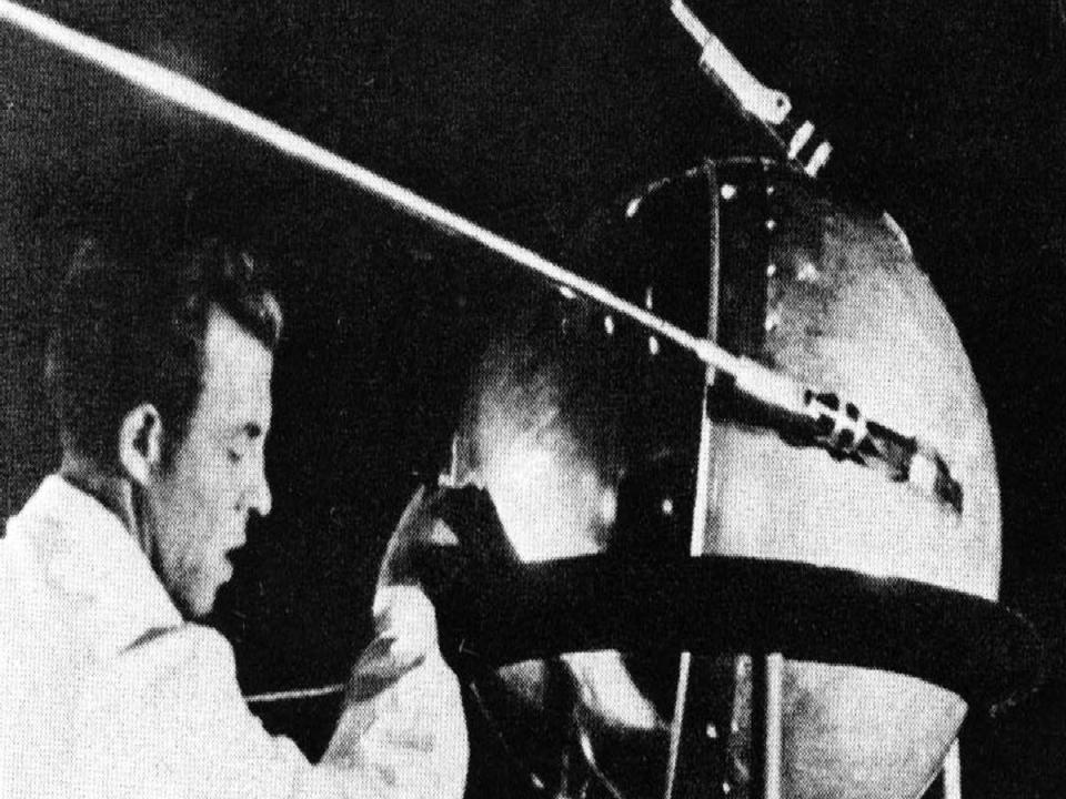 a black and white photo shows a man in a lab coat in front of the sputnik satellite