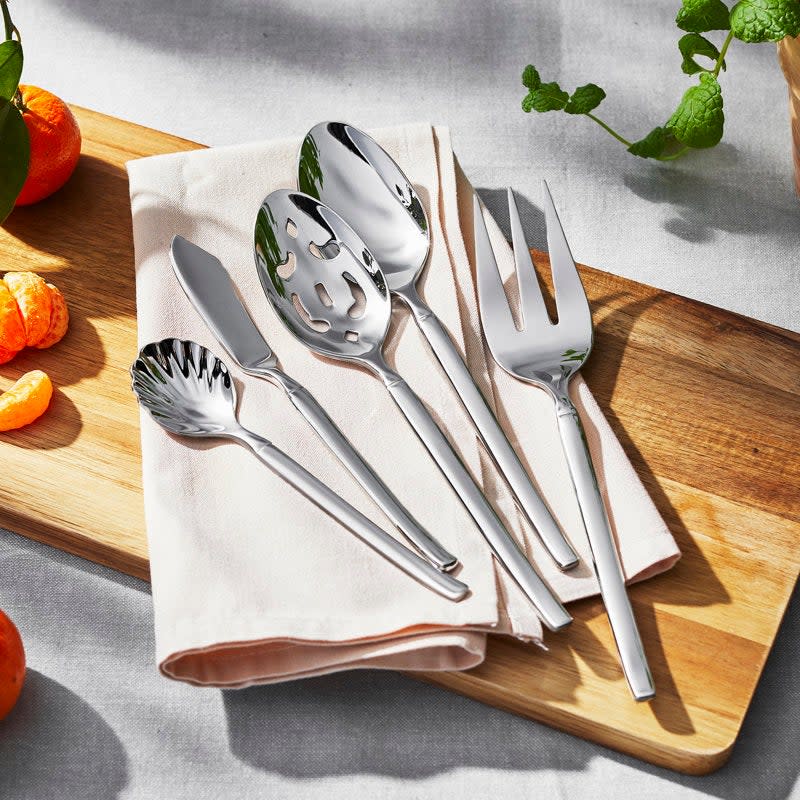Silverware set including a spoon, fork, slotted spoon, and server on a wooden board next to a napkin