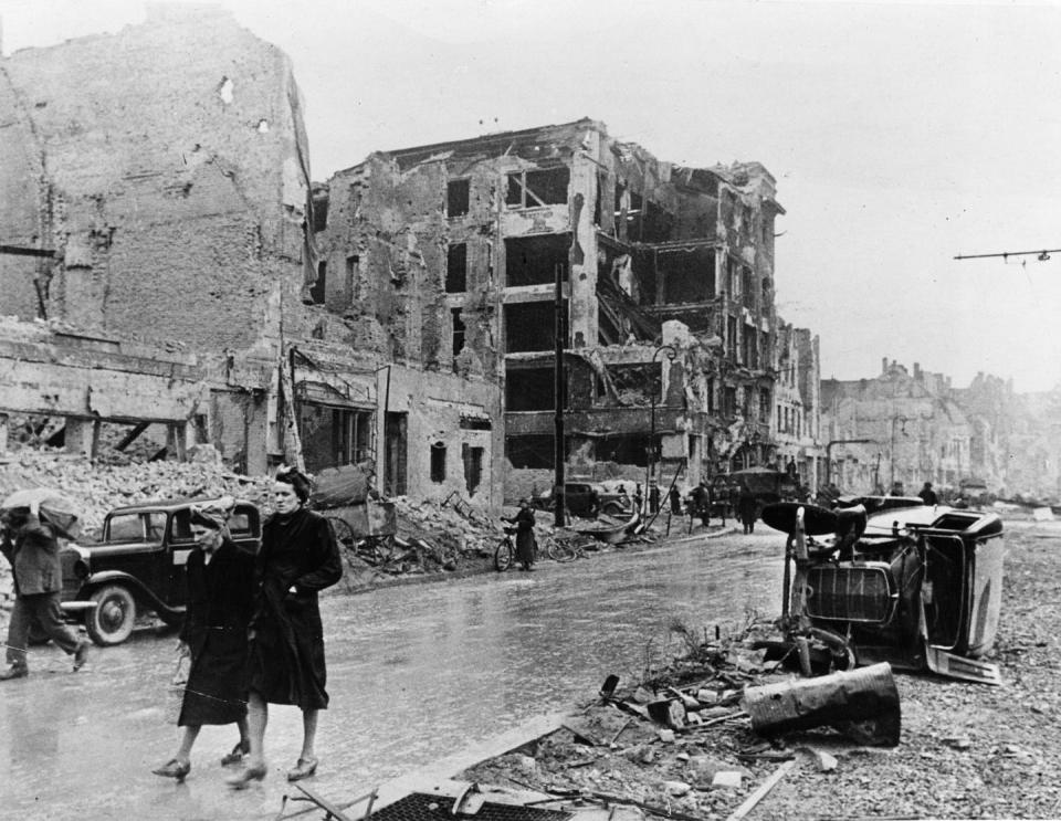 Two women are walking on a street with an overturned car and devastated building in the background.