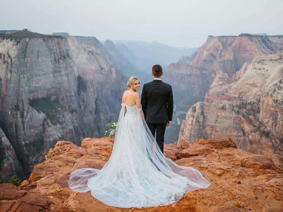 A bride looks over her shoulder while holding hands with her groom on top of a mountain.