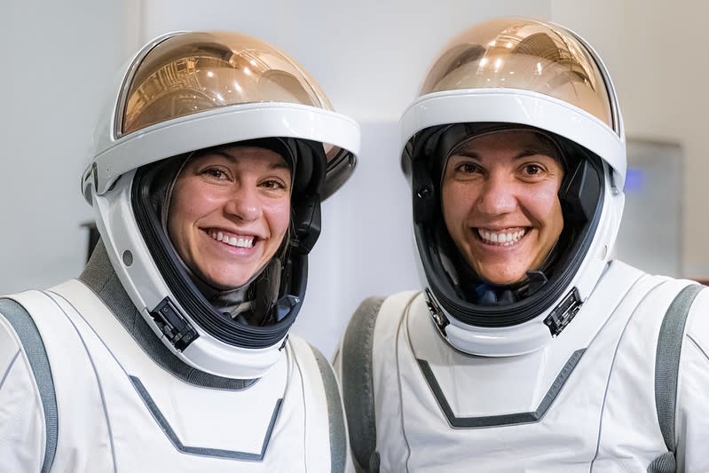 Two women in white spacesuits, seen from the chest up. Their copper-colored visors are up, their faces padded inside their helmets. They are in a white room, smiling.