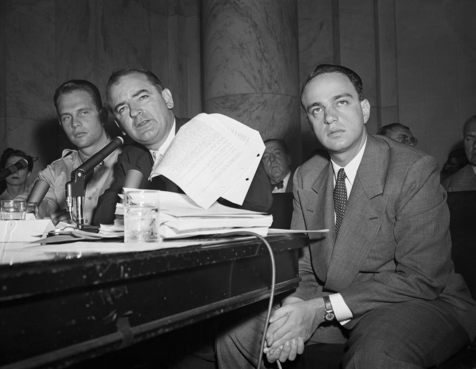 joseph mccarthy flipping through papers as he speaks into a microphone while roy cohn sits in a chair next to him with his hands calsped