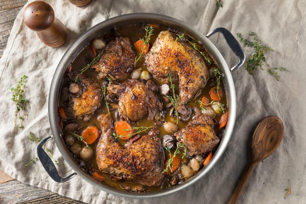 overhead view of pot of homemade french coq au vin chicken with veggies and sauce on white cloth with sprigs of herbs and a wooden spoon