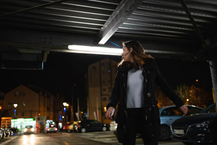 A woman wearing a coat walks under a metal structure in a dimly lit parking lot at night with buildings and parked cars in the background