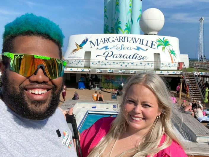 مردی با موهای آبی، عینک آفتابی سبز و پیراهن خاکستری و زنی با موهای بلوند و لباس صورتی در یک کشتی با تابلویی که روی آن نوشته شده است «Margaritaville Sea Paradise» سلفی می گیرند.