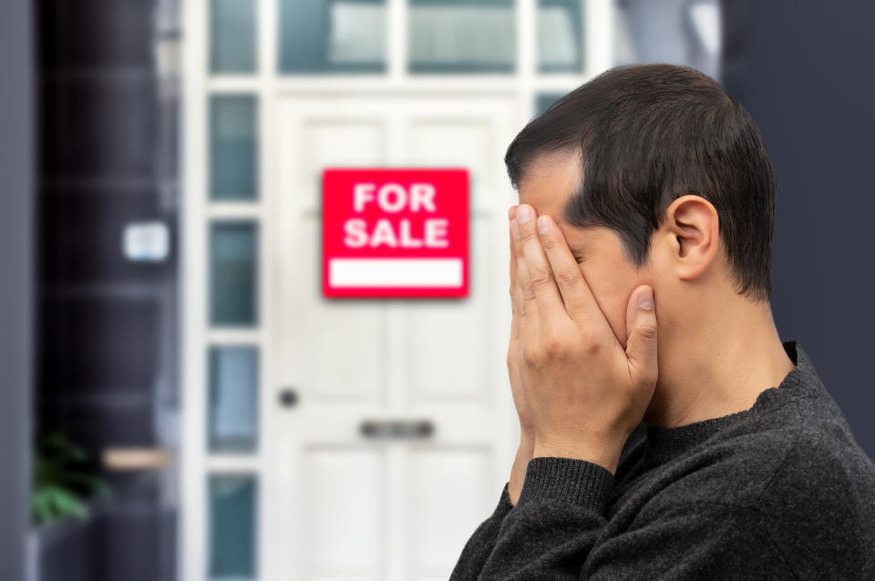 A man covering his face with his hands stands outside a house with a "For Sale" sign on it